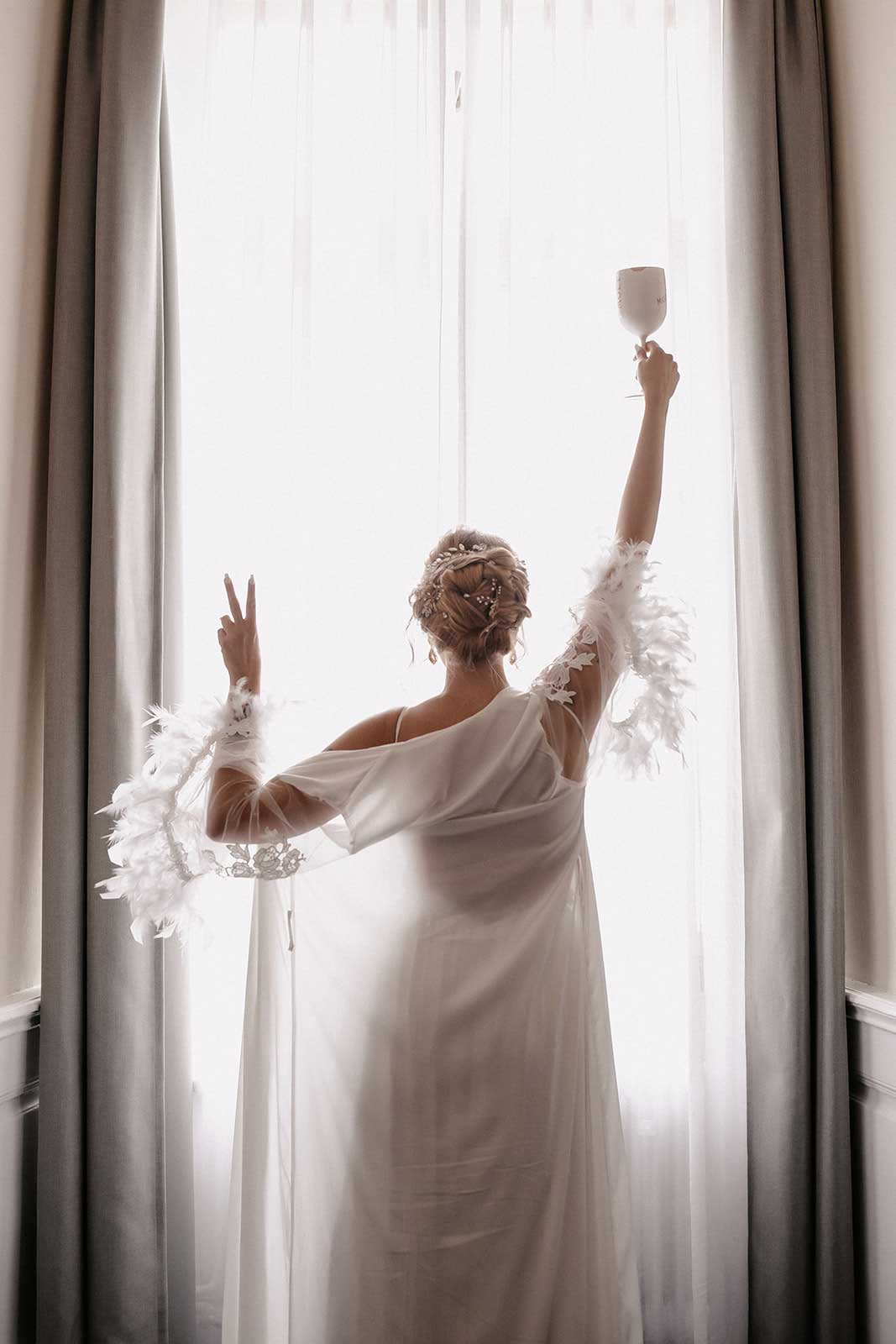 getting ready Hochzeitsfotograf Berlin un der Braut im Federkleid prostet am Fenster dem Tag zu.