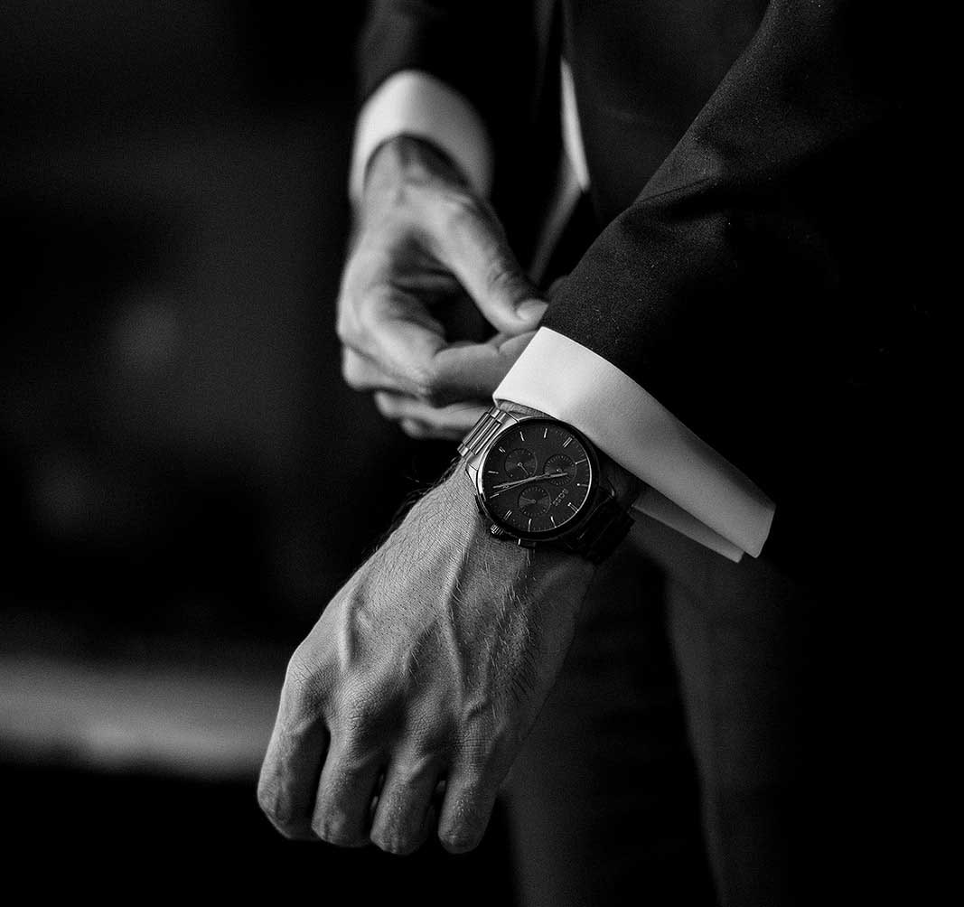 Detailaufnahme der Hand eines Bräutigams, der eine stilvolle Uhr mit schwarzem Zifferblatt justiert, während er sich auf seine Hochzeit vorbereitet