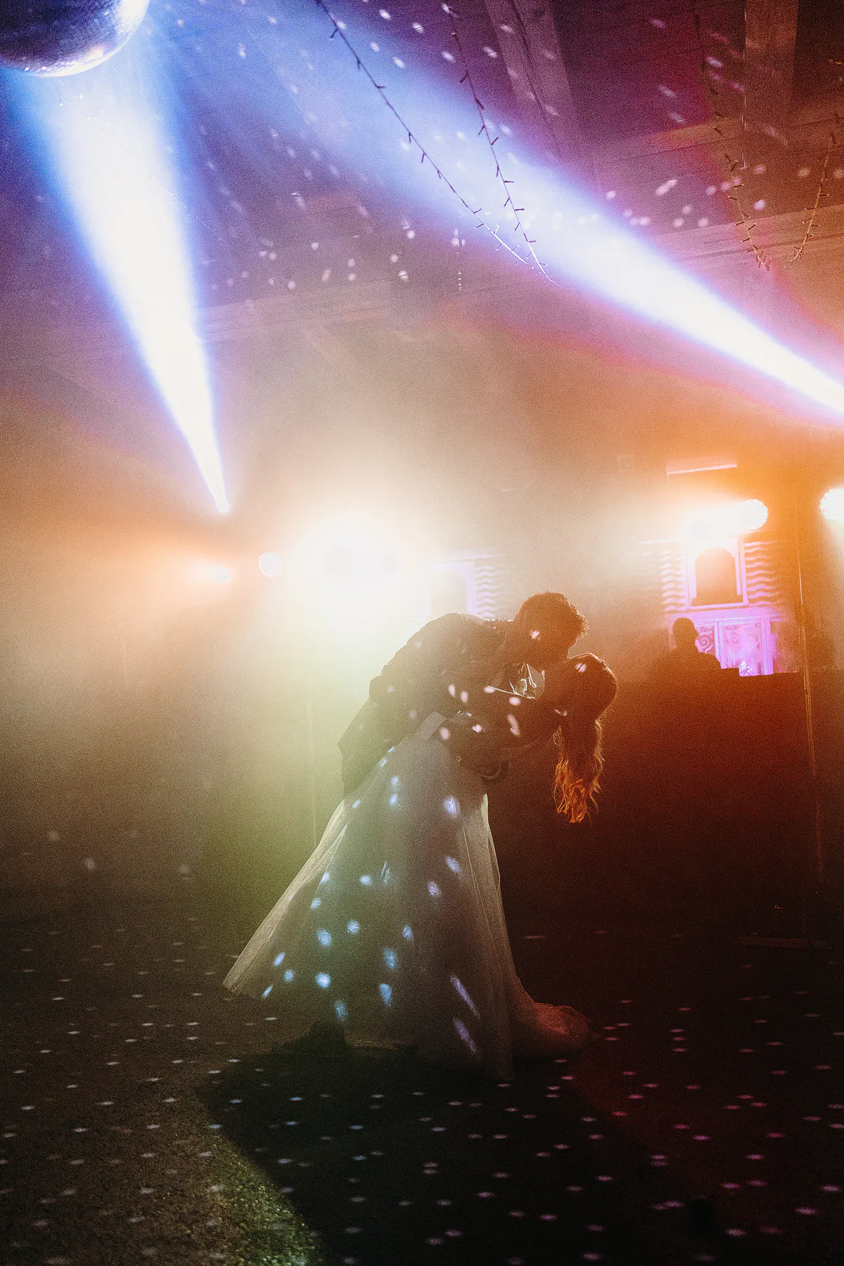 Ein Hochzeitspaar küsst sich tief unter farbigen Lichtstrahlen und Diskokugelreflexen
