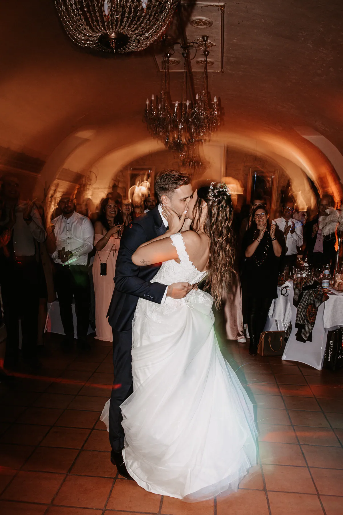 Ein Hochzeitspaar küsst sich beim Tanz unter Kronleuchtern, umringt von Gästen