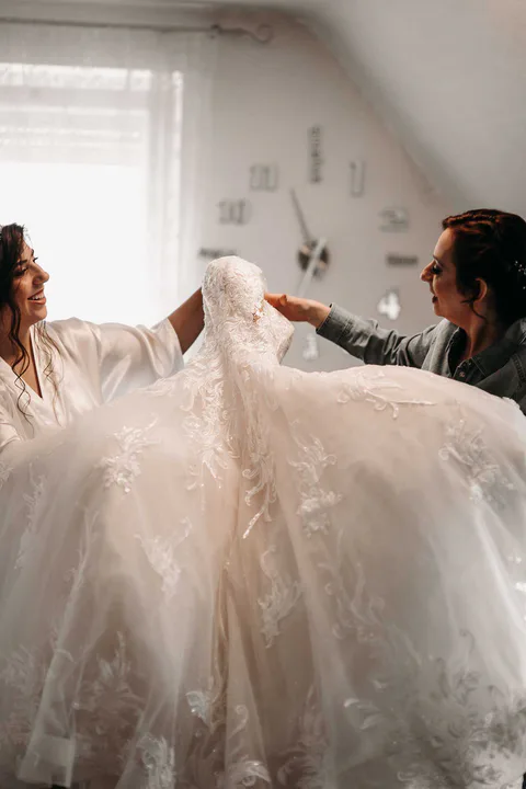 Zwei Personen halten ein Brautkleid vor der Braut hoch