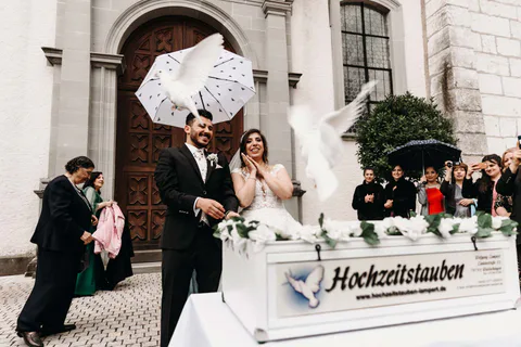 Zwei weiße Tauben fliegen vor dem Brautpaar und einer Kirche